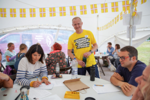 Alan in the Comms Unplugged tent with bright yellow not worrier / warrior message emblazoned on the front. Unpluggers at a table getting involved in the activity. 