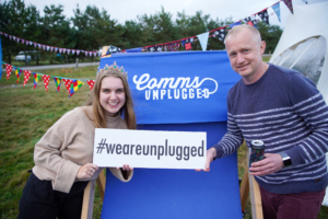 Lauren and Alan sharing a smile at Comms Unplugged. Tent and bunting in the background holding a blue #weareunplugged sign board. 