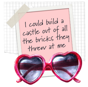 red heart shaped sunglasses with a piece of paper in pink taped to a board with the wording 'I could build a castle out of all the bricks they threw at me'
